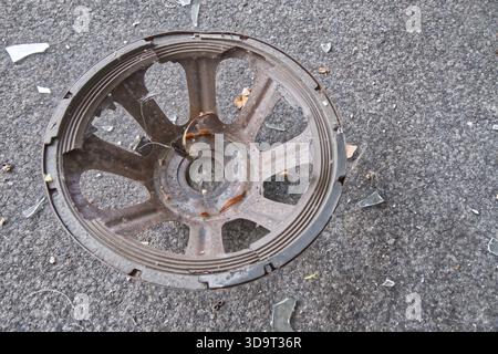 A rusty, damaged metal wheel rim lying on a gray surface, surrounded by small pieces of glass and debris. Stock Photo