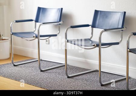 Three modern blue chairs with chrome frames arranged in a minimalist interior setting, placed on a textured gray rug against a white wall. Stock Photo