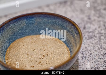 A close-up view of a shallow bowl filled with brown sugar, resting on a textured countertop. The bowl has a blue and brown gradient design, highlighti Stock Photo