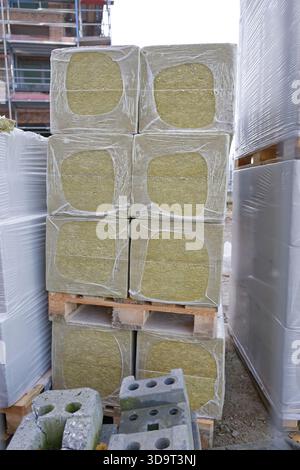 A stack of rectangular bales wrapped in plastic, arranged on wooden pallets at a construction site. Surrounding materials include concrete blocks and Stock Photo