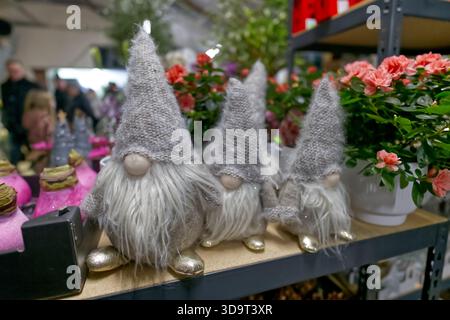 Three decorative gnomes with gray hats and white beards, sitting on a shelf among potted flowers and plants in a vibrant market setting. Stock Photo