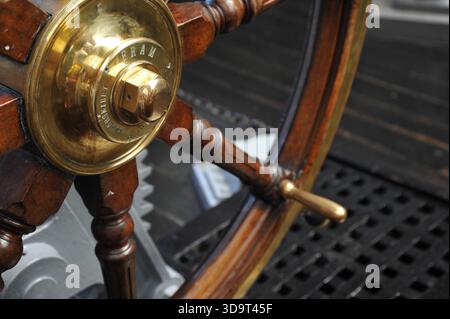A close-up view of a vintage ship's steering wheel, showcasing its wooden structure and brass components. The wheel is mounted on a metal base, with a Stock Photo