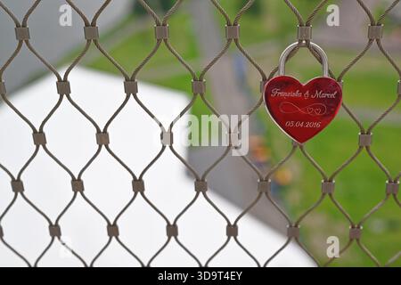 A red heart-shaped padlock hangs on a metal mesh fence, engraved with names and a date. The background features a blurred green landscape, suggesting Stock Photo