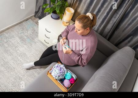 Woman sitting on a gray sofa crocheting with yarn and needle supplies. for illustrating home hobbies, relaxation, crafting, and handmade content. Stock Photo