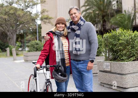 Senior couple posing on urban walkway with road bicycle and hanging helmet wearing scarf and beanie Stock Photo
