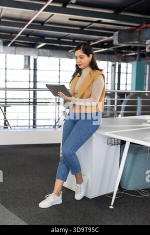 Mid adult woman wearing business attire perching on edge of white desk holding tablet in workspace Stock Photo