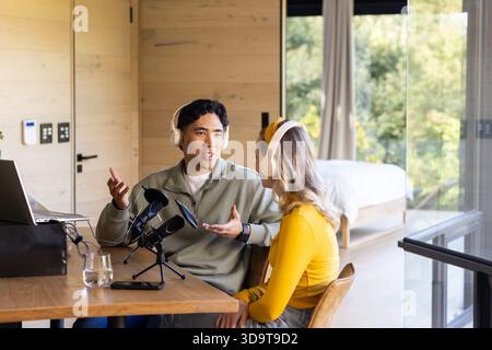 Diverse friends recording podcast at wooden table in home studio with microphone, headphones Stock Photo