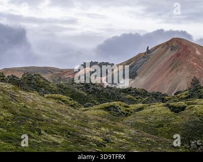 Mossy lava rocks and steaming geothermal ground near a towering, colorful rhyolite mountain spire. Stock Photo