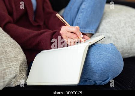 Woman sitting cross-legged on dark fabric sofa writing in open blank notebook with rose gold pen Stock Photo