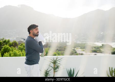 Indian man standing at terrace railing wiping towel holding water bottle by succulents, copy space Stock Photo