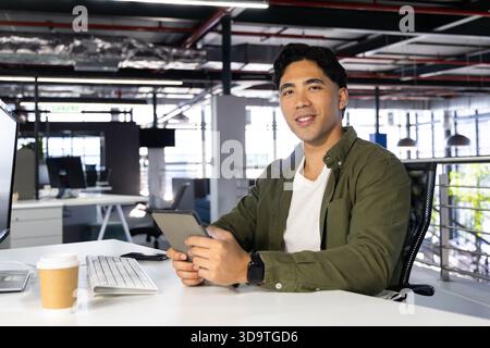 Asian adult male sitting at office holding tablet checking smartphone beside keyboard coffee cup Stock Photo