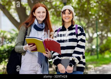 Diverse female students studying on campus walkway holding folder and tablet, wearing backpacks Stock Photo
