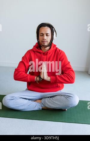 Middle aged man meditating cross-legged on green yoga mat at home wearing white over-ear headphones Stock Photo