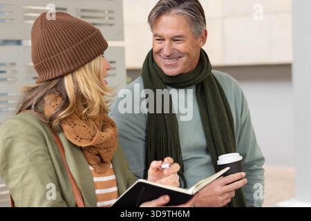 Couple leaning against metal bench in urban plaza, comparing notes in notebook and pen over coffee Stock Photo