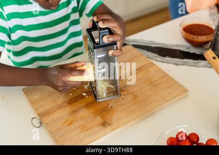 Boy shredding cheese on box grater at kitchen counter with tomato sauce, pizza pan, cherry tomatoes Stock Photo