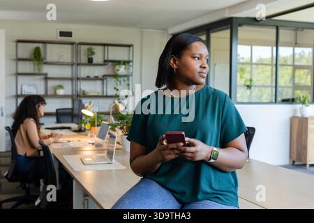 Female coworkers sitting at shared desk, one holding smartphone, other working on laptop Stock Photo