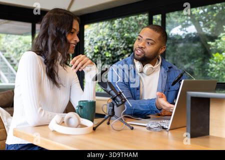 Diverse podcast hosts sitting at table by glass windows using laptop and speaking into mics Stock Photo