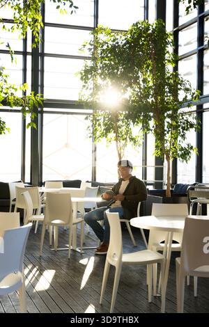 Asian man sitting and using smartphone in modern lounge atrium amid white tables and potted trees Stock Photo