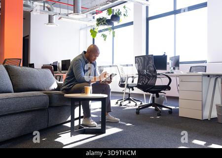 African American man sitting on sofa in open-plan office holding tablet and coffee cup, copy space Stock Photo