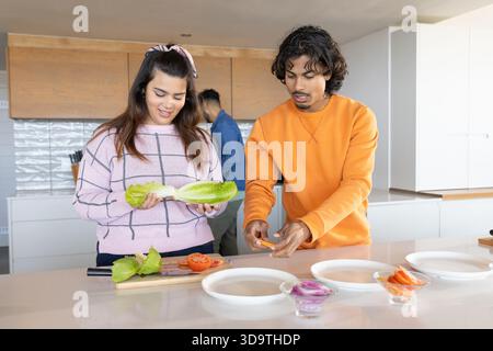 Friends prepping at kitchen island, woman holding romaine in pink check sweater, man slicing pepper Stock Photo