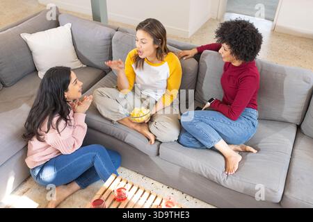 Three females talking, sharing potato chips on gray sectional sofa at home with red drinks Stock Photo