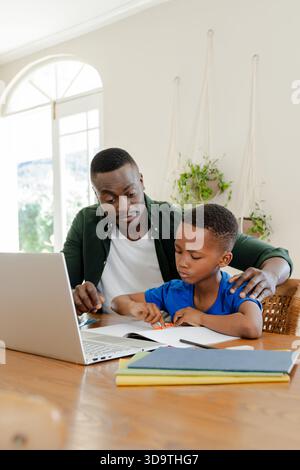African American father and son sitting at wooden dining table working on open workbook with laptop Stock Photo