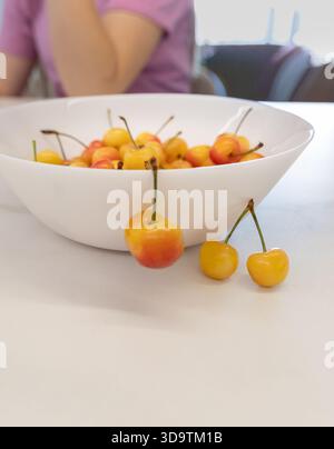 Close up shot of a white bowl filled with yellow and red cherries on a white table with two cherries outside the bowl, with soft focus background Stock Photo