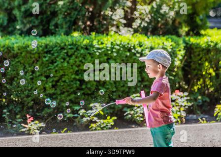 Child playing with bubble wand in Bacău, Romania, surrounded by floating soap bubbles and greenery. Stock Photo
