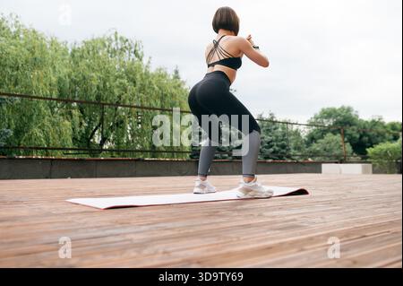 Strong sportswoman doing sit ups on fitness mat on wooden area. Back view of beautiful girl doing exercise in sportswear and sneakers. Concept of spor Stock Photo
