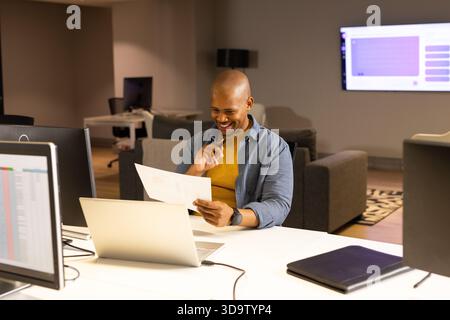 African American man reviewing document at office desk while viewing spreadsheet on laptop monitor Stock Photo