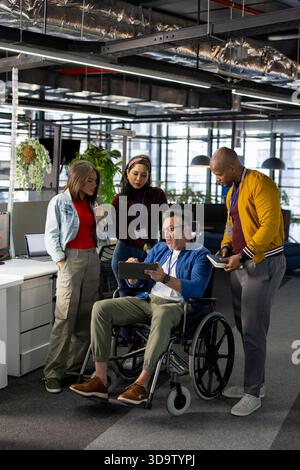 Diverse coworkers collaborating at office around man in wheelchair holding tablet and smartphone Stock Photo