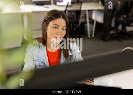 Indian woman wearing denim jacket holding chart and analyzing data at office desk with computer Stock Photo