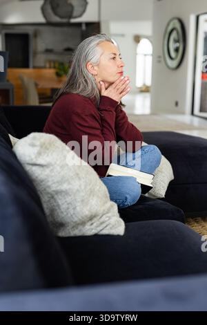Senior female sitting on dark-blue sofa in living room holding open book and gazing toward mirror Stock Photo
