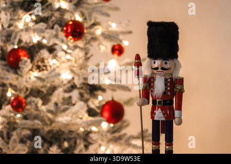 Wooden nutcracker toy standing next to a decorated Christmas tree with warm lights and red ornaments. Stock Photo
