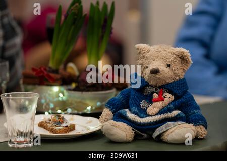 Cute teddy bear in a knitted sweater sitting on a festive holiday table setting. Adorable plush toy bear surrounded by cozy Christmas décor. Stock Photo