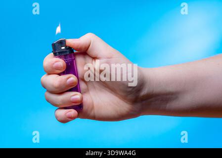 A close-up of a child's hand holding a lit lighter against a plain blue background. Fire safety concept. Stock Photo