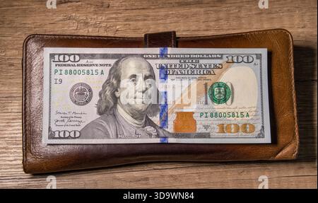 Brown leather wallet with a crisp $100 bill featuring Benjamin Franklin, placed on a wooden surface. Stock Photo