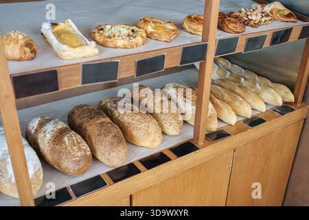 Variety of fresh baked sourdough, seeded bread, croissants, danishes, and baguettes display on wooden shelf in bakery shop Stock Photo