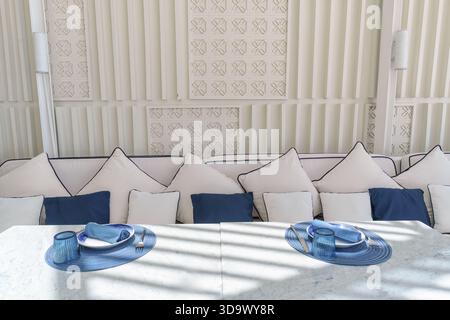 Mediterranean restaurant interior featuring blue and white dining table, tableware, cushion seat, sunlight on modern wall Stock Photo