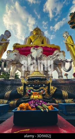 Elaborate Buddhist Monument Featuring Large Gold Deity, Attendant Statues, and Traditional Spirit Offerings Under a Blue Sky Stock Photo