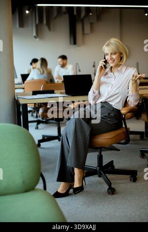 A businesswoman in formal attire talks on her phone while seated in a contemporary office. Others are focused on their work in the background, creatin Stock Photo