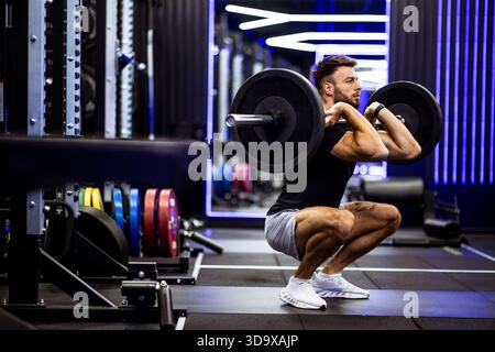 A man performs a barbell squat in a contemporary gym with vibrant lighting. He focuses intently on his workout, surrounded by various weight plates an Stock Photo