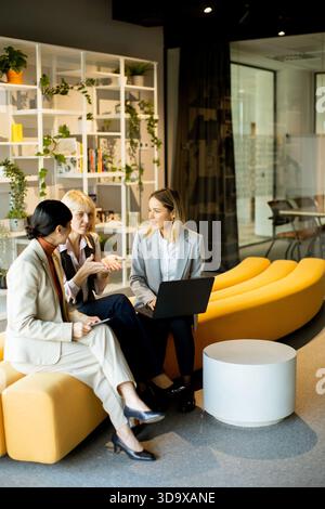 In a stylish co-working area, three women engage in a discussion while seated on a curved yellow sofa. One woman uses a laptop while the others contri Stock Photo