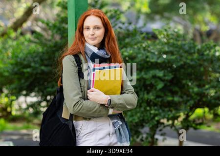 White adult female student standing on campus path carrying backpack holding colorful notebooks Stock Photo