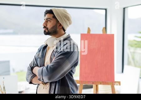 Indian male standing by studio window wearing headphones, gazing at lake with red canvas, easel Stock Photo