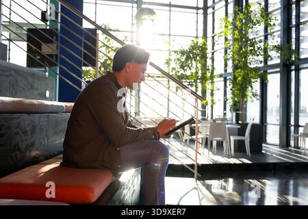 Man sketching on tablet with stylus while sitting on cushioned bench by metal staircase and windows Stock Photo
