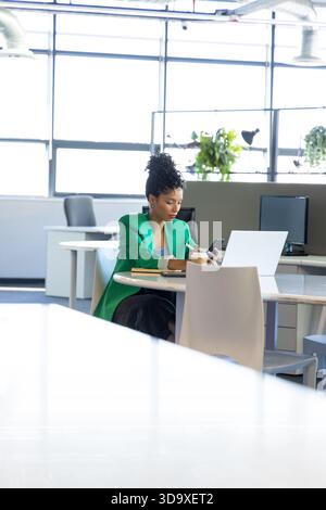 African American woman checking smartphone and jotting notes in notebook at open-plan office Stock Photo