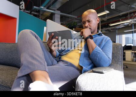 African American man reclining on sofa in office holding tablet supporting chin near smartphone Stock Photo
