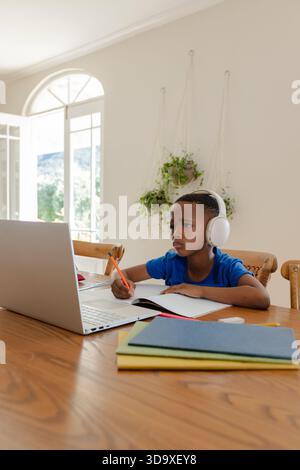 African American child sitting at wooden dining table at home, working on laptop with orange pencil Stock Photo