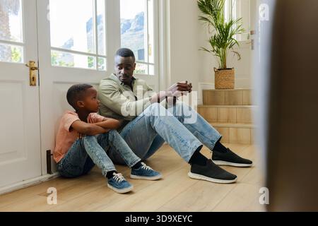 African American father and son sitting on light wood foyer floor, talking beside glass doors Stock Photo